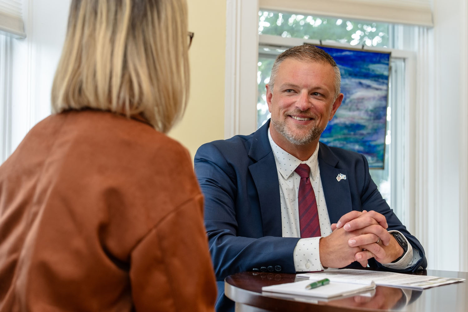 A man in a suit and tie sits and smiles while in a meeting with a woman with short blonde hair.