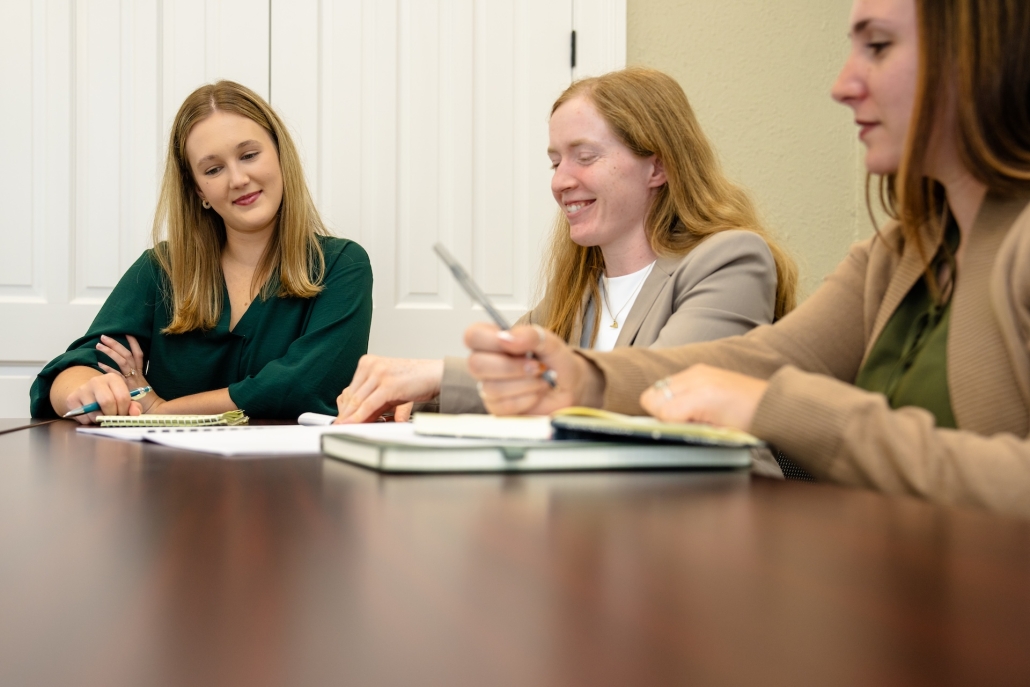 Three women sit at a table in a meeting and review paperwork.